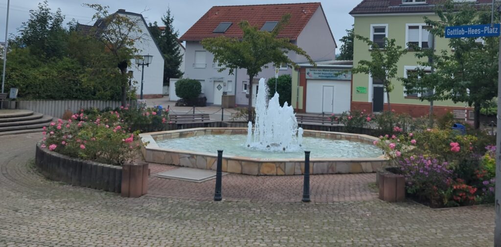 Ein Springbrunnen mit gepflasterten Platz drum herum. Vereinzelt Blumen und Sitzbänke