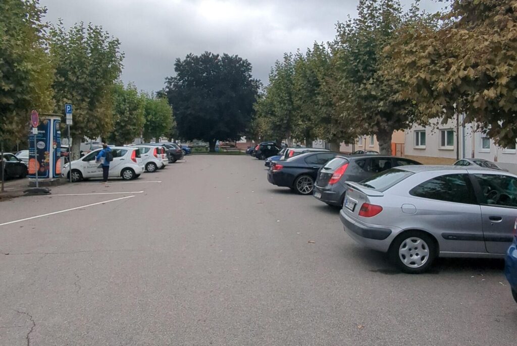 Ein großer Parkplatz aus Beton mit vielen Autos. Um dem Parkplatz stehen einzelne Bäume.