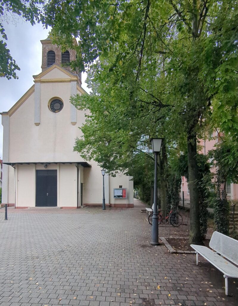 Eine Kirche an einem zugepflasterten Platz. Rechts stehen Bäume in einer Reihe. Ein begrünter Weg ist neben der Kirche erkennbar.